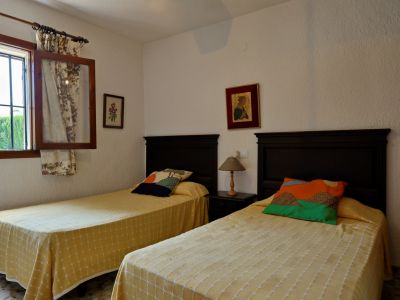 Dormitorio doble con cabeceros de madera oscura, colchas amarillas estampadas y cojines coloridos. Ventana con rejas y cortinas florales.
