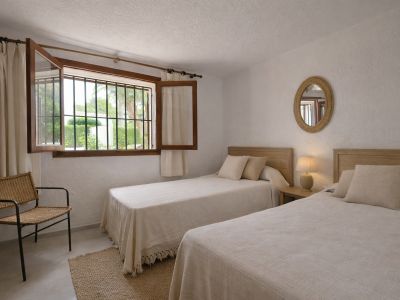 Dormitorio luminoso con dos camas individuales, cabeceros de madera clara y ventana abierta con rejas que ofrece vistas al jardín. Incluye mesita de noche, lámpara y silla de mimbre.