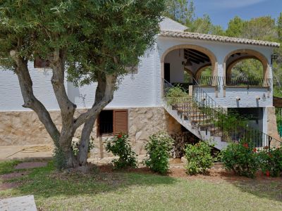 El exterior de la villa muestra una fachada blanca texturizada sobre una base de piedra, con una terraza cubierta de arcos y una escalera exterior de azulejos, junto a un olivo.