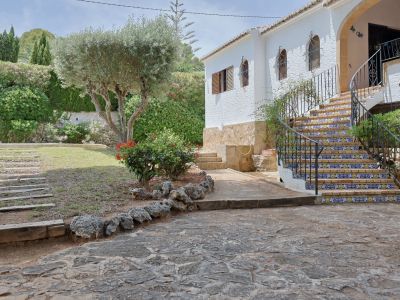 Entrada principal de la villa con escalera curva de azulejos decorativos y barandilla de hierro forjado, flanqueada por un jardín mediterráneo con olivo y camino de piedra.