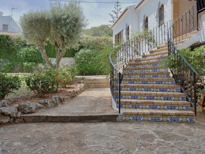 Escalera exterior de ladrillo con azulejos decorativos azules y amarillos, flanqueada por un jardín mediterráneo con olivo y camino de piedra.
