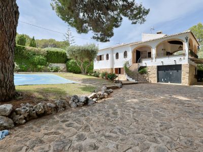 Exterior de la villa con amplia entrada empedrada, destacando la fachada blanca con arcos y la escalera principal. Se aprecia la zona de piscina cubierta y vegetación mediterránea.