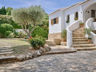 Fachada blanca de la villa con escalera curva de piedra que conduce a la entrada. El jardín mediterráneo presenta un olivo central, arbustos y flores, con un camino de losas.
