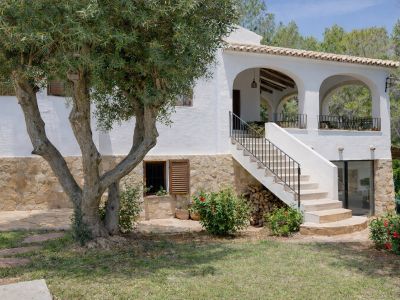 Fachada de villa con base de piedra y muros blancos, destacando un olivo maduro y una escalera exterior que conduce a una terraza porticada con arcos y barandilla de hierro.