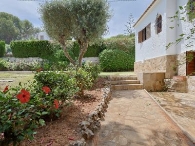 Lateral de la villa con fachada blanca y zócalo de piedra. Un patio de losas conduce a escalones de piedra, rodeado por un jardín con hibiscos rojos y un olivo.