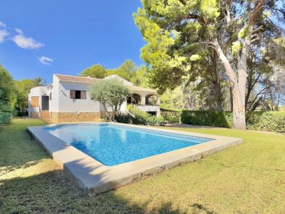 Piscina rectangular con azulejos azules, rodeada de césped verde bien cuidado y una villa blanca con terraza cubierta, enmarcada por pinos y olivos.
