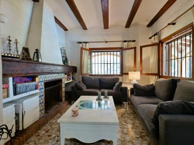 Salón con chimenea de obra y azulejos decorativos, vigas de madera vistas y suelo de terrazo. Ventanas de madera con rejas aportan luz natural.