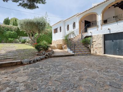 Vista exterior de la villa con entrada empedrada, escalera curva de azulejos mosaico y barandilla de hierro forjado. Un jardín mediterráneo con olivo y césped complementa la fachada blanca.