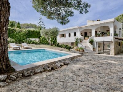 Vista exterior de una villa blanca tradicional con piscina privada, patio empedrado y escalera exterior. Rodeada de un exuberante jardín mediterráneo.