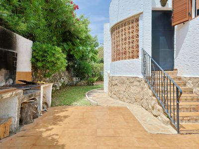Zona de barbacoa exterior con terraza de baldosas, escalera con barandilla de hierro forjado y fachada de estuco blanco con celosía decorativa, rodeada de vegetación mediterránea.