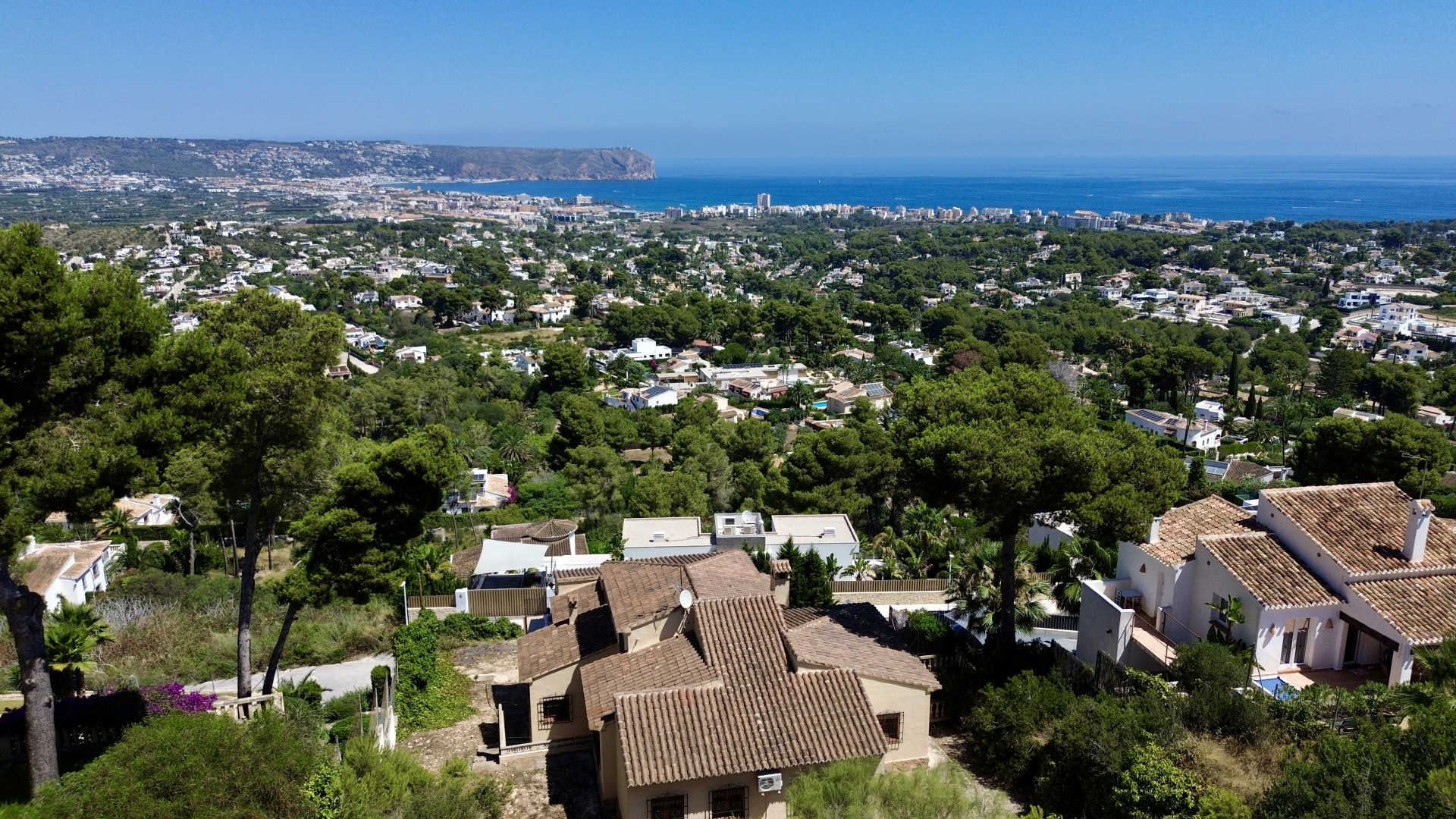 Vista aérea de Jávea, mostrando villas con tejados de teja entre pinos, con el mar Mediterráneo y el Cabo de San Antonio al fondo. Vista aérea de Jávea, mostrando villas con tejados de teja entre pinos, con el mar Mediterráneo y el Cabo de San Antonio al fondo.
