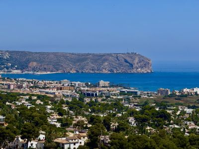 Amplia vista panorámica que muestra el paisaje residencial de Jávea, el azul intenso del Mediterráneo con barcos y el prominente cabo rocoso con su faro.