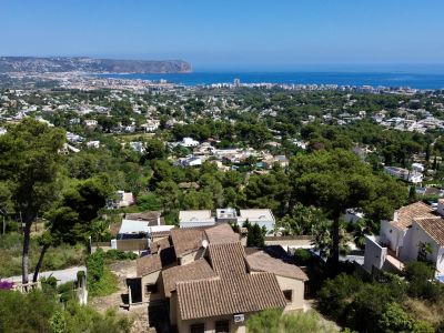 Vista aérea de Jávea, mostrando villas con tejados de teja entre pinos, con el mar Mediterráneo y el Cabo de San Antonio al fondo.