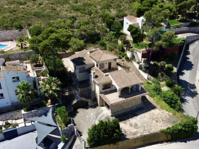 Vista aérea de la villa de varios niveles con tejados tradicionales, múltiples terrazas y áreas ajardinadas en pendiente, integrada en la vegetación mediterránea.