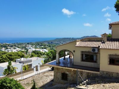 Vista exterior de la villa con terraza cubierta de diseño arqueado y barandales negros. Se aprecian vistas panorámicas al mar, la ciudad y la montaña desde la parcela en pendiente.