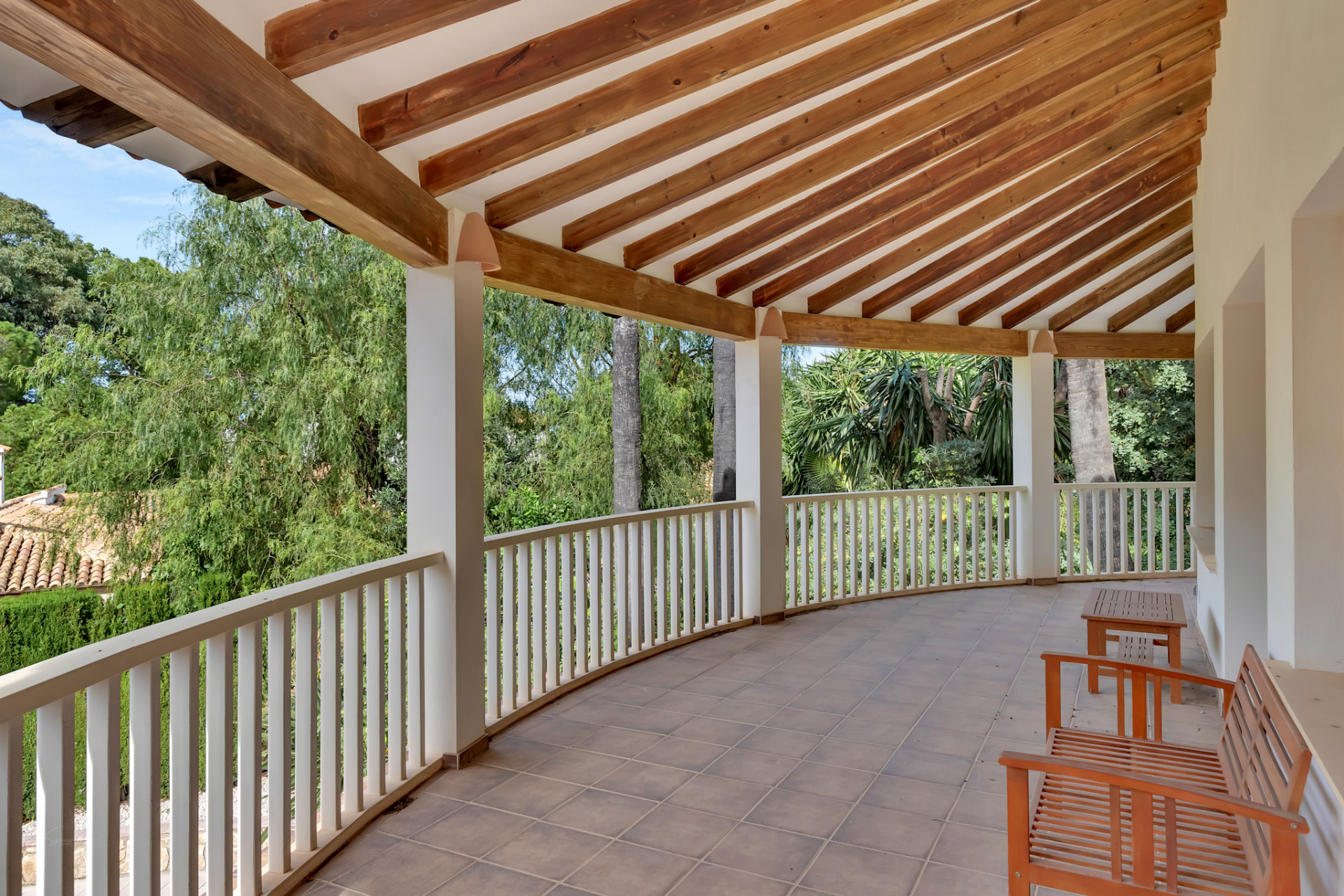 Amplia terraza cubierta de forma curva con suelo de baldosas y un techo de vigas de madera vistas. La barandilla blanca ofrece vistas a la exuberante vegetación, con mobiliario de madera. Amplia terraza cubierta de forma curva con suelo de baldosas y un techo de vigas de madera vistas. La barandilla blanca ofrece vistas a la exuberante vegetación, con mobiliario de madera.