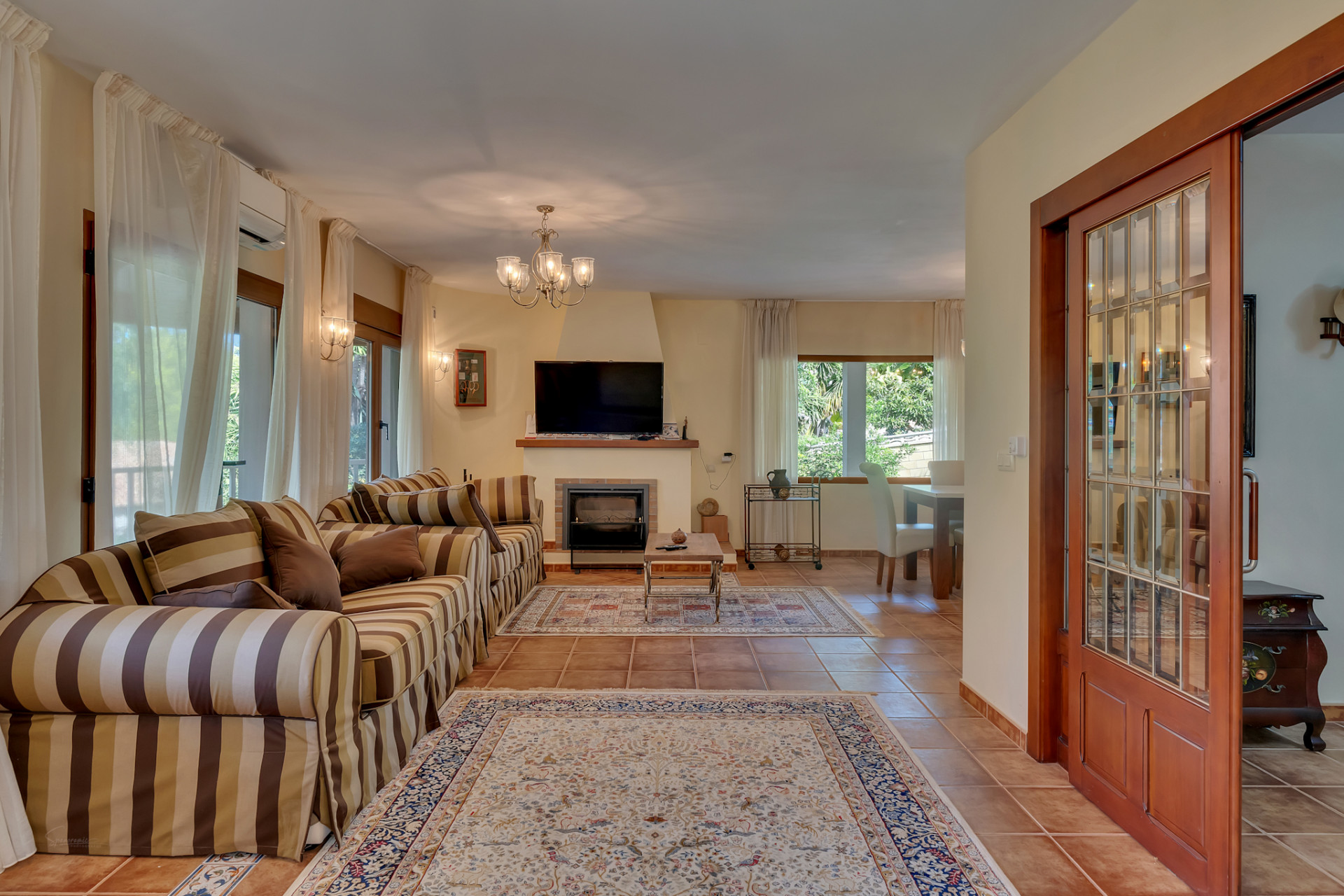 Amplio salón con sofá a rayas, chimenea y televisión. Destacan los ventanales con cortinas claras, el suelo de terracota y la puerta corredera de madera. Amplio salón con sofá a rayas, chimenea y televisión. Destacan los ventanales con cortinas claras, el suelo de terracota y la puerta corredera de madera.