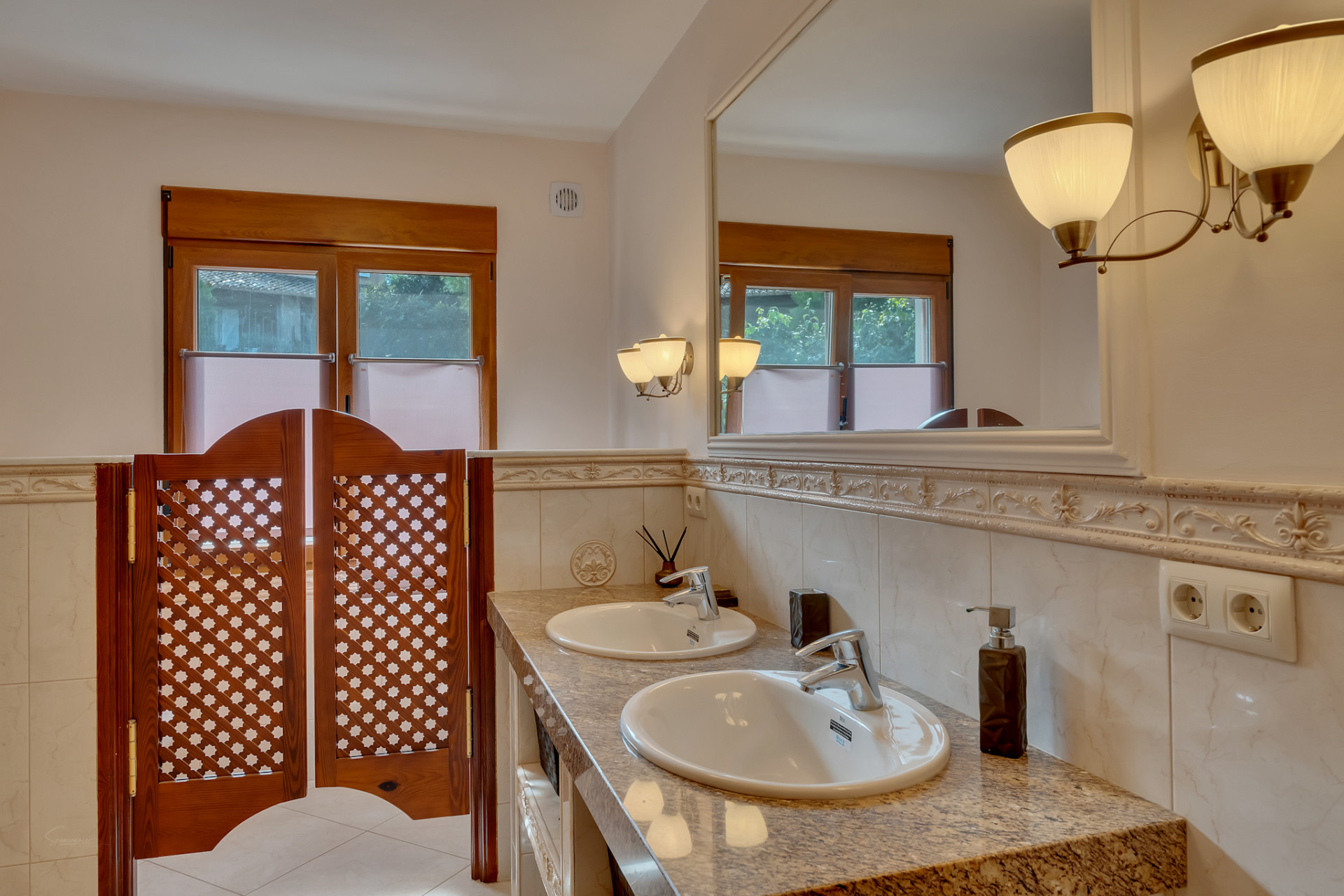 Baño con doble lavabo sobre encimera de granito, gran espejo enmarcado y apliques de luz. Ventanas de madera y puertas tipo saloon con celosía. Baño con doble lavabo sobre encimera de granito, gran espejo enmarcado y apliques de luz. Ventanas de madera y puertas tipo saloon con celosía.