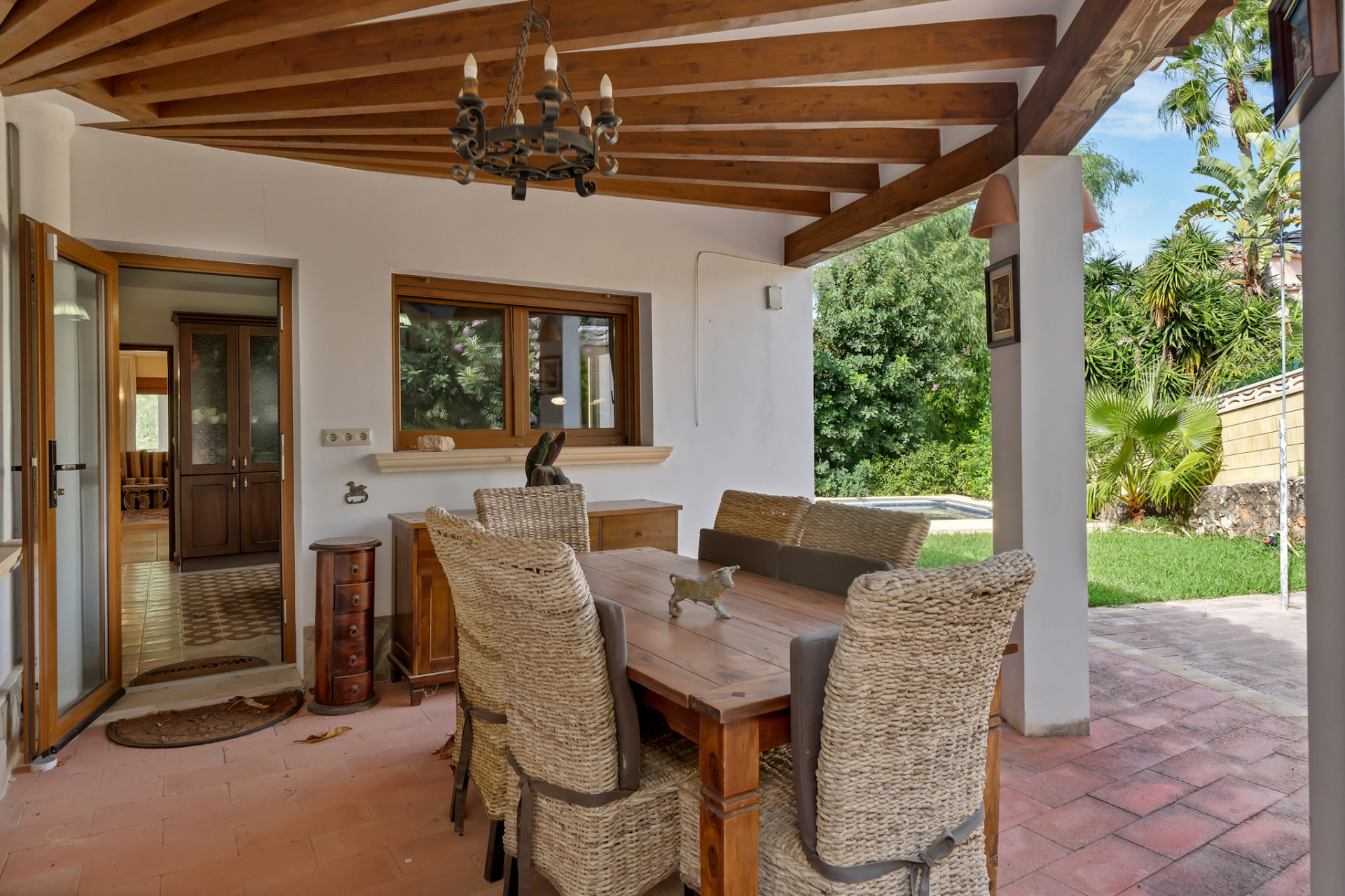 Comedor exterior cubierto con techo de vigas de madera, suelo de terracota y lámpara rústica de forja. Sillas de mimbre rodean una mesa de madera, con vistas al jardín y acceso interior. Comedor exterior cubierto con techo de vigas de madera, suelo de terracota y lámpara rústica de forja. Sillas de mimbre rodean una mesa de madera, con vistas al jardín y acceso interior.