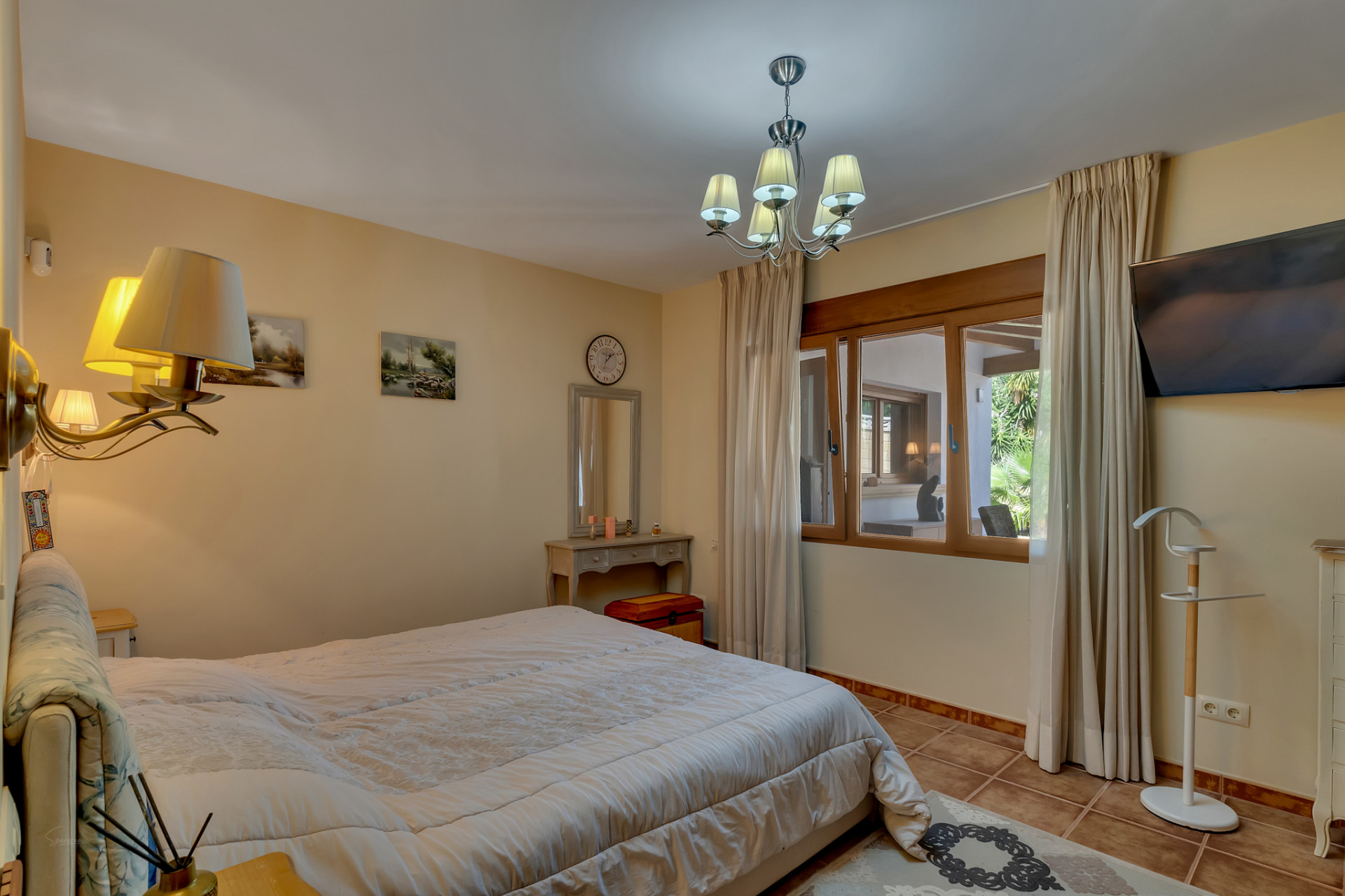 Dormitorio principal con cama grande, suelo de baldosas y ventana con marco de madera que ofrece vistas a una zona cubierta y al jardín. Incluye tocador y TV. Dormitorio principal con cama grande, suelo de baldosas y ventana con marco de madera que ofrece vistas a una zona cubierta y al jardín. Incluye tocador y TV.