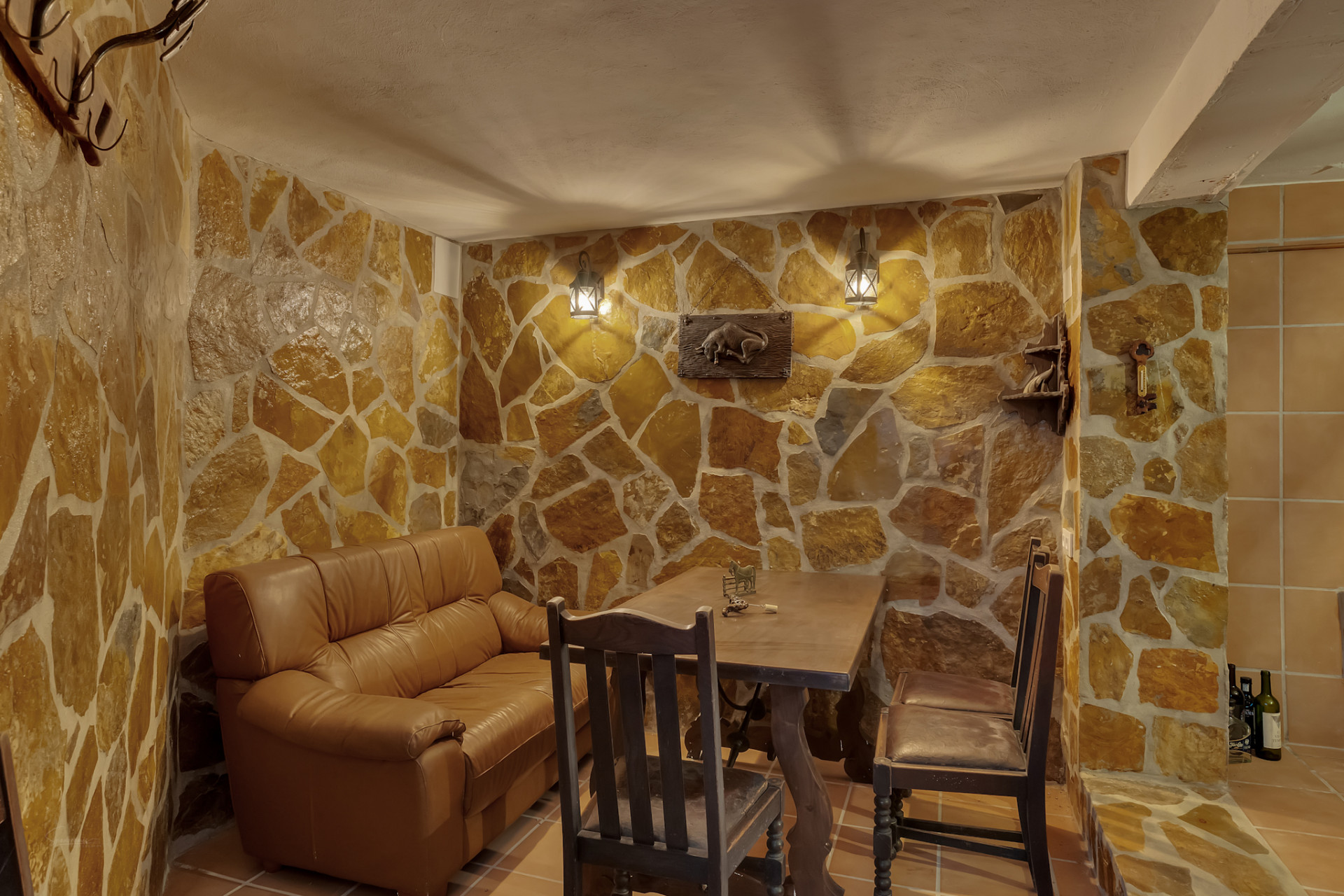 Espacio interior con paredes revestidas de piedra irregular y suelo de baldosas. Incluye un sofá de cuero, mesa rústica de madera y cálida iluminación de faroles. Espacio interior con paredes revestidas de piedra irregular y suelo de baldosas. Incluye un sofá de cuero, mesa rústica de madera y cálida iluminación de faroles.