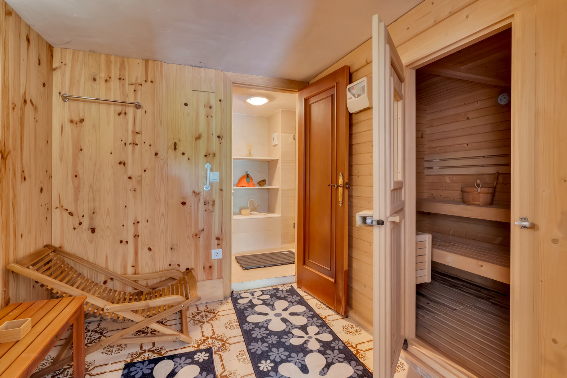 Zona de relajación con paredes de madera clara, suelo de baldosas y acceso a una sauna de pino. Incluye una silla de madera y una entrada a un área de servicio con estantes. Zona de relajación con paredes de madera clara, suelo de baldosas y acceso a una sauna de pino. Incluye una silla de madera y una entrada a un área de servicio con estantes.