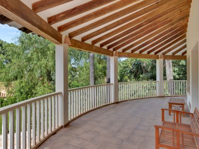 Amplia terraza cubierta de forma curva con suelo de baldosas y un techo de vigas de madera vistas. La barandilla blanca ofrece vistas a la exuberante vegetación, con mobiliario de madera.