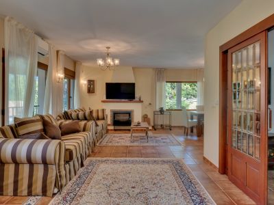 Amplio salón con sofá a rayas, chimenea y televisión. Destacan los ventanales con cortinas claras, el suelo de terracota y la puerta corredera de madera.