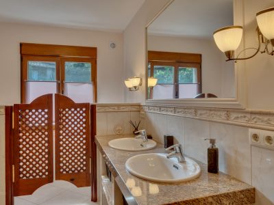 Baño con doble lavabo sobre encimera de granito, gran espejo enmarcado y apliques de luz. Ventanas de madera y puertas tipo saloon con celosía.