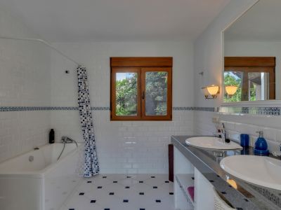 Baño con doble lavabo sobre encimera oscura y espejo grande. Azulejos blancos tipo metro con cenefa azul decorativa, suelo estampado y ventana de madera con vistas a la vegetación.