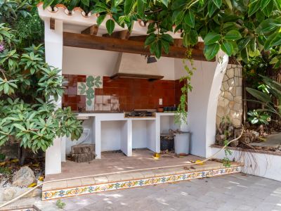 Cocina exterior con barbacoa integrada, salpicadero de azulejos terracota con motivo de cactus y techo de vigas de madera. El área tiene suelo de baldosas y está rodeada de vegetación.
