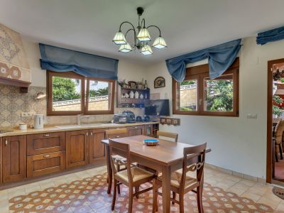 Cocina funcional con muebles de madera oscura, azulejos decorativos y suelo de baldosas estampadas. Dos ventanas con vistas al jardín y una mesa de comedor de madera.