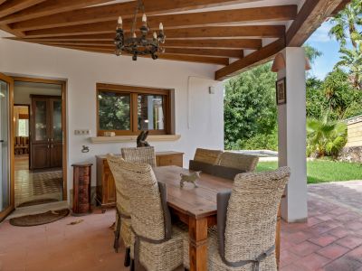 Comedor exterior cubierto con techo de vigas de madera, suelo de terracota y lámpara rústica de forja. Sillas de mimbre rodean una mesa de madera, con vistas al jardín y acceso interior.