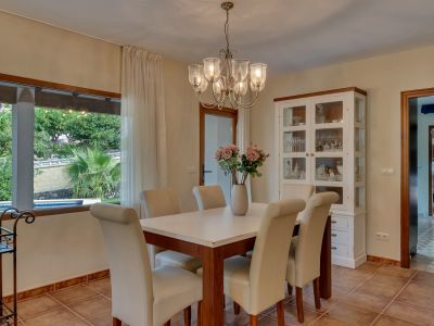 Comedor luminoso con mesa de madera, sillas tapizadas en crema y lámpara de araña. Amplio ventanal con vistas al exuberante jardín y piscina privada.