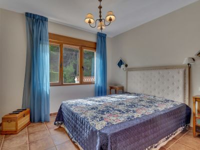 Dormitorio con cama de cabecero capitoné blanco, colcha azul estampada y suelo de baldosas de terracota. Ventana de madera con cortinas azules ofrece luz natural y vistas a la vegetación.