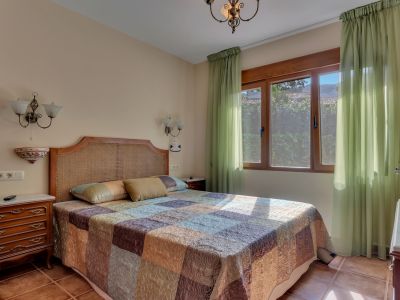 Dormitorio luminoso con cabecero de ratán, colcha estampada y una gran ventana de madera con cortinas verdes translúcidas. Suelo de baldosas de terracota y mesitas de noche clásicas de madera.