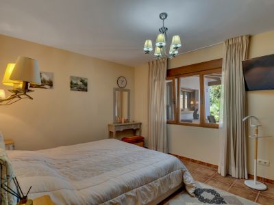 Dormitorio principal con cama grande, suelo de baldosas y ventana con marco de madera que ofrece vistas a una zona cubierta y al jardín. Incluye tocador y TV.