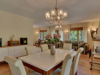 Elegante comedor con araña clásica y sillas color crema, conectado a una luminosa sala de estar con sofás a rayas y grandes ventanales con vistas a la vegetación.