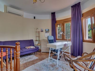 Luminosa oficina con sofá morado, escritorio de madera clara y silla ergonómica. Grandes ventanas de madera con vistas a la vegetación, suelo de baldosas de terracota y aire acondicionado.