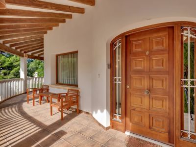 Terraza de entrada cubierta con puerta de madera panelada, suelo de baldosas y vigas de madera expuestas. Incluye zona de asientos y barandilla curva con vistas verdes.