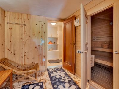 Zona de relajación con paredes de madera clara, suelo de baldosas y acceso a una sauna de pino. Incluye una silla de madera y una entrada a un área de servicio con estantes.