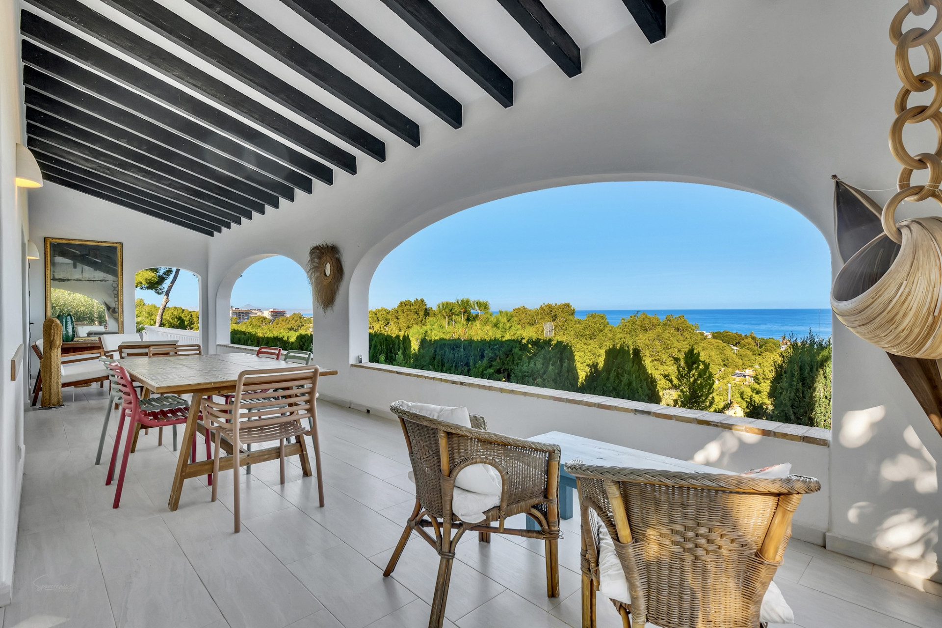 Amplia terraza cubierta con vigas de madera oscura y arcos, ofreciendo vistas panorámicas a la vegetación y al mar Mediterráneo. Incluye comedor y zona de asientos de mimbre. Amplia terraza cubierta con vigas de madera oscura y arcos, ofreciendo vistas panorámicas a la vegetación y al mar Mediterráneo. Incluye comedor y zona de asientos de mimbre.
