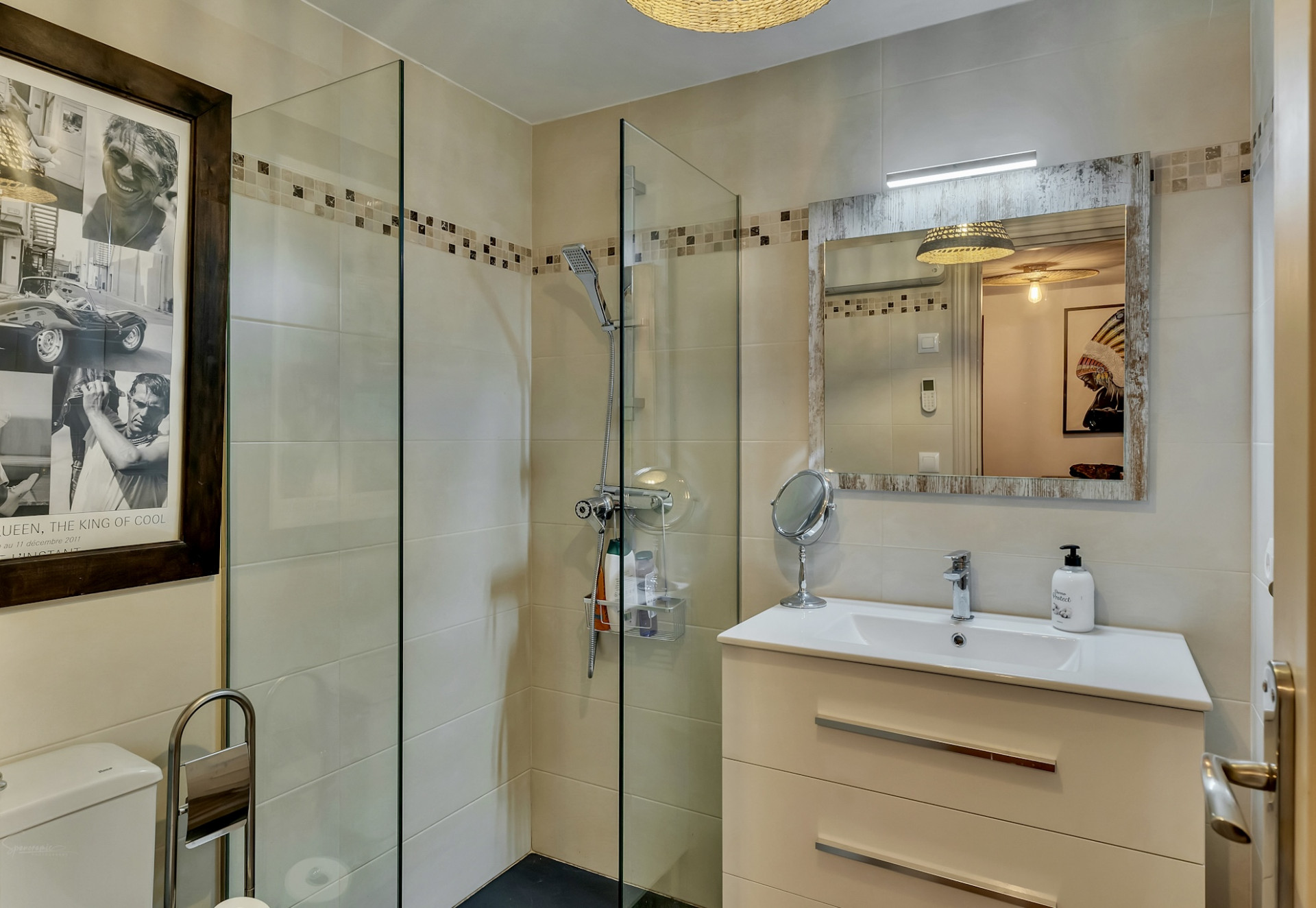 Baño moderno con ducha de cristal, azulejos beige y cenefa decorativa. Incluye un mueble de lavabo blanco con espejo de marco rústico y luz integrada. Baño moderno con ducha de cristal, azulejos beige y cenefa decorativa. Incluye un mueble de lavabo blanco con espejo de marco rústico y luz integrada.