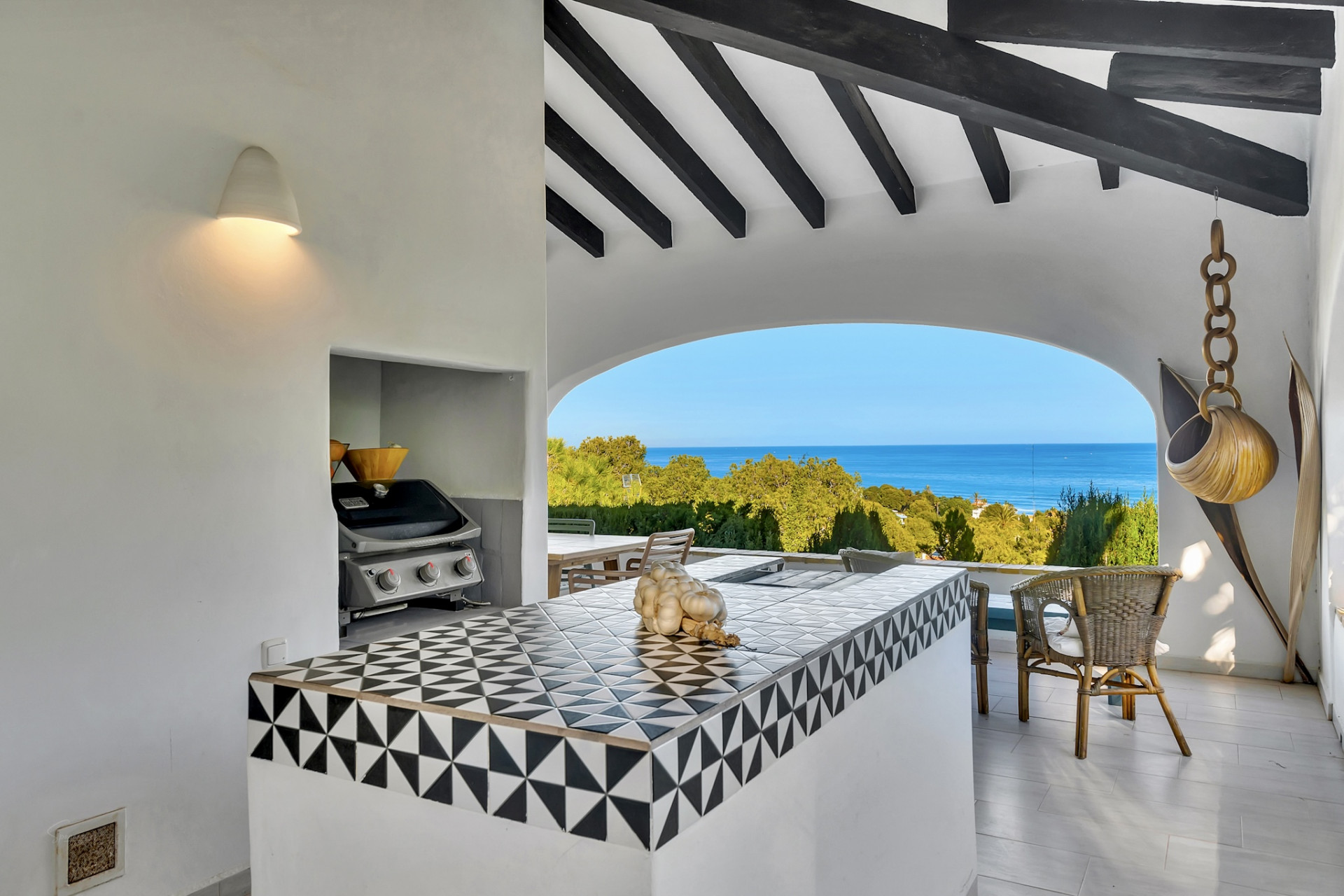 Cocina exterior bajo pérgola con encimera de azulejos geométricos en blanco y negro, barbacoa integrada y vistas panorámicas al mar Mediterráneo a través de un arco. Cocina exterior bajo pérgola con encimera de azulejos geométricos en blanco y negro, barbacoa integrada y vistas panorámicas al mar Mediterráneo a través de un arco.