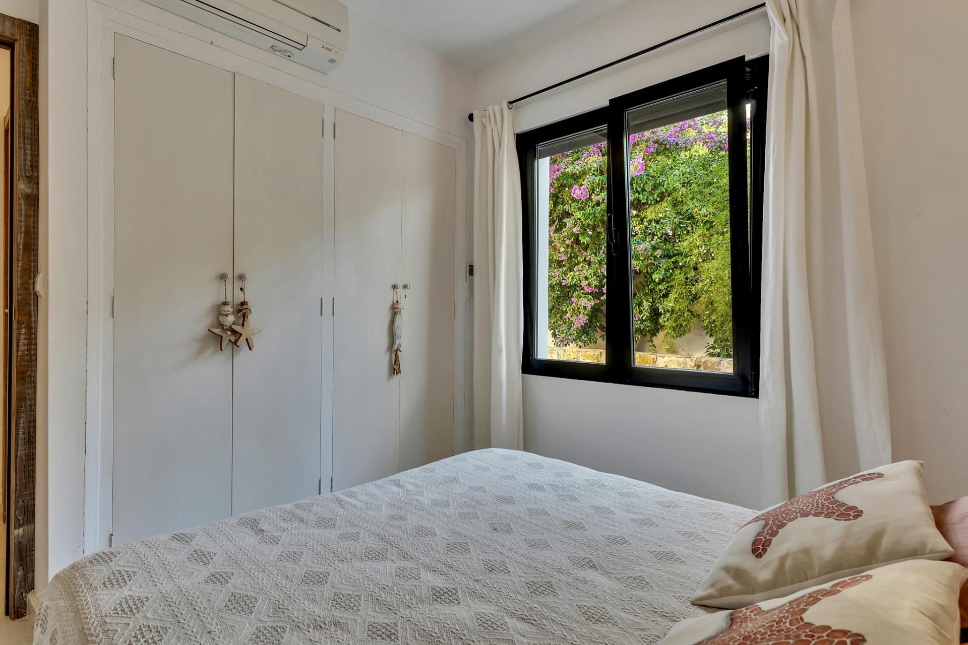 Dormitorio con armarios empotrados blancos, unidad de aire acondicionado y ventana que enmarca una vista vibrante de buganvillas rosadas. Dormitorio con armarios empotrados blancos, unidad de aire acondicionado y ventana que enmarca una vista vibrante de buganvillas rosadas.