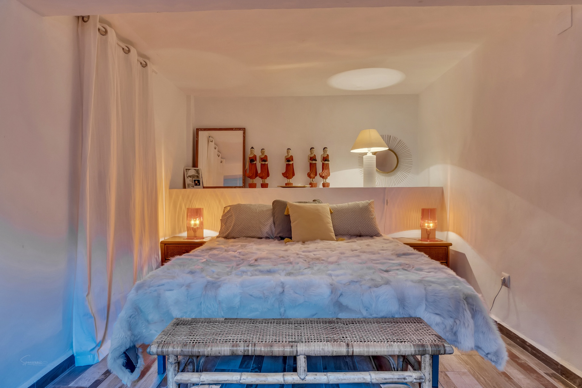 Dormitorio con cama de diseño, cabecero con estante decorado con figuras y lámparas. Banco de ratán y cortinas blancas complementan el ambiente cálido. Dormitorio con cama de diseño, cabecero con estante decorado con figuras y lámparas. Banco de ratán y cortinas blancas complementan el ambiente cálido.