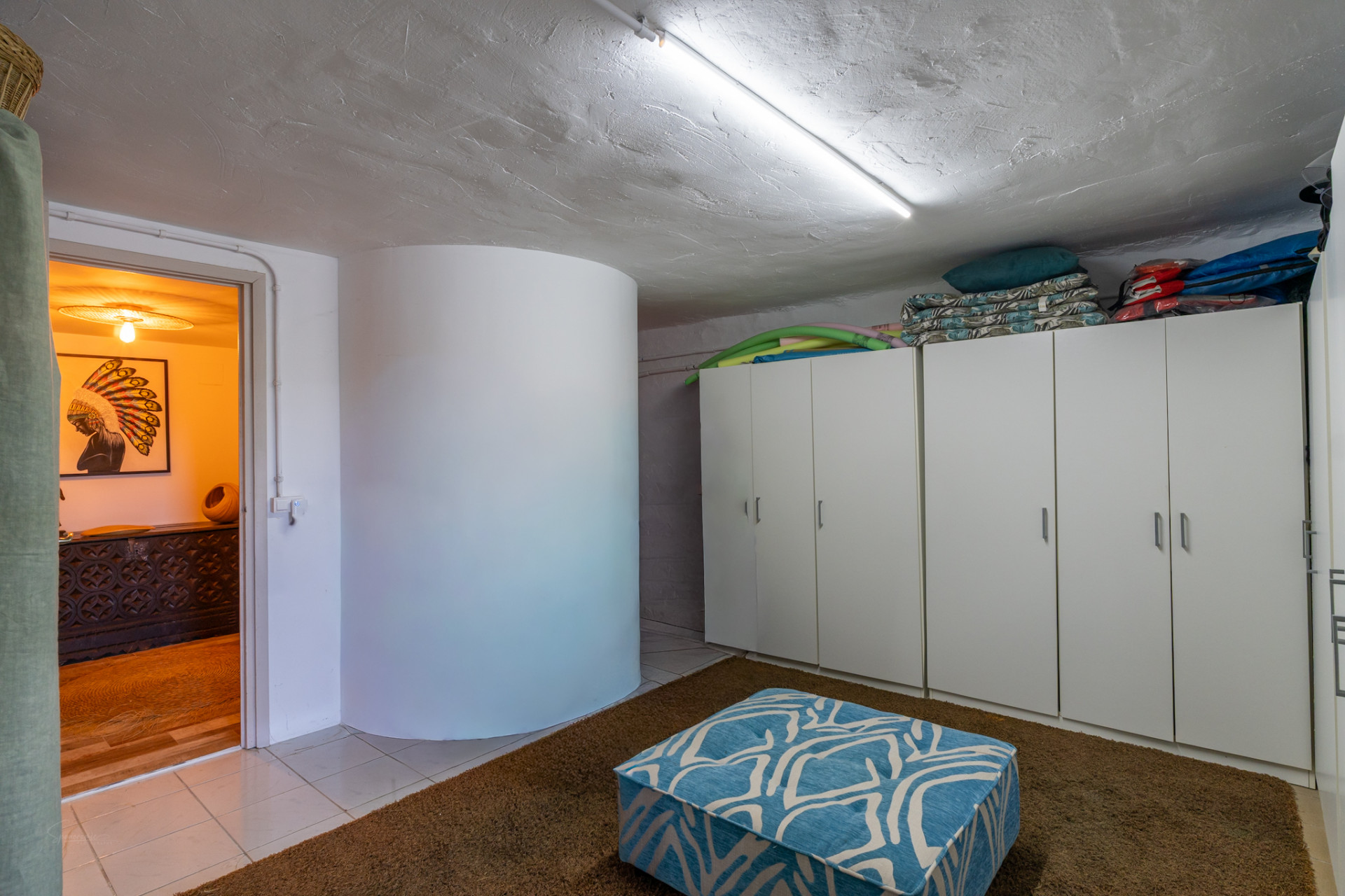 Espacio práctico con armarios blancos para almacenamiento, techo texturizado con luz fluorescente y alfombra marrón. Un puf azul estampado y una puerta abierta revelan la cálida decoración de una sala adyacente. Espacio práctico con armarios blancos para almacenamiento, techo texturizado con luz fluorescente y alfombra marrón. Un puf azul estampado y una puerta abierta revelan la cálida decoración de una sala adyacente.