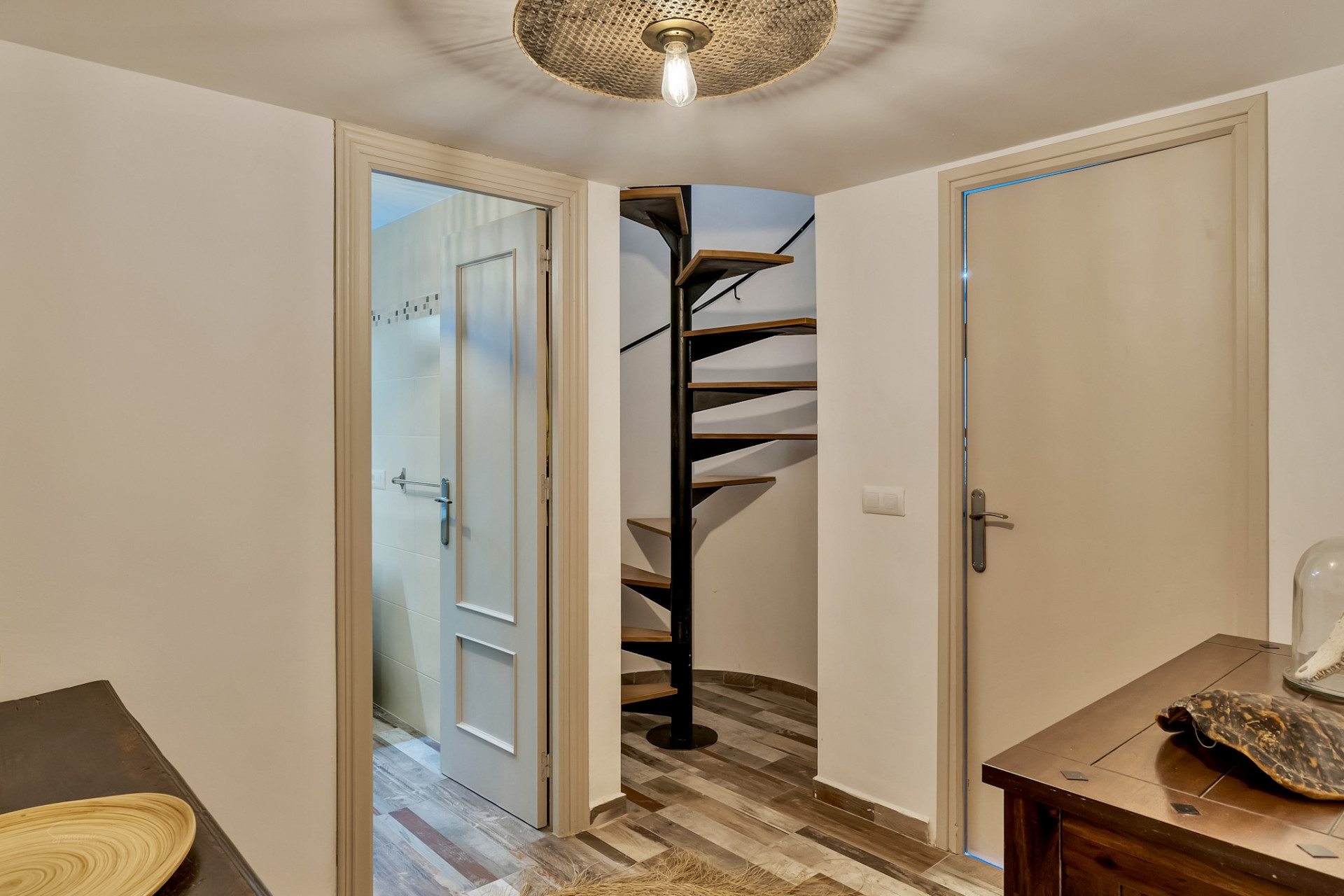 Pasillo con una distintiva escalera de caracol de madera y metal negro, complementado por un suelo de baldosas efecto madera. Se observan dos puertas, una de ellas abierta a un baño, y una consola de madera oscura. Pasillo con una distintiva escalera de caracol de madera y metal negro, complementado por un suelo de baldosas efecto madera. Se observan dos puertas, una de ellas abierta a un baño, y una consola de madera oscura.