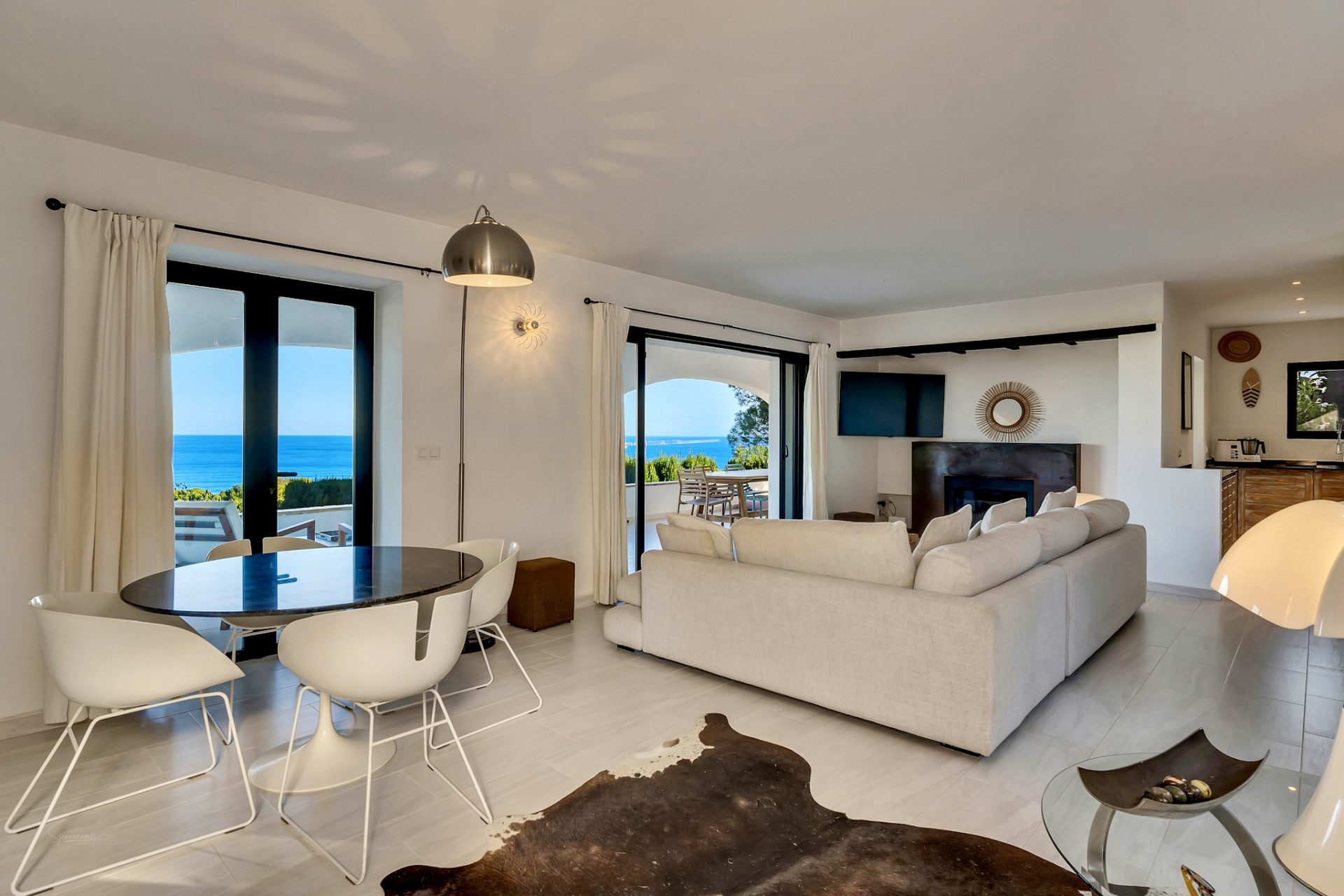 Salón comedor de planta abierta con mesa ovalada oscura, sillas blancas y sofá claro, que se abre a una terraza con vistas panorámicas al mar. Salón comedor de planta abierta con mesa ovalada oscura, sillas blancas y sofá claro, que se abre a una terraza con vistas panorámicas al mar.