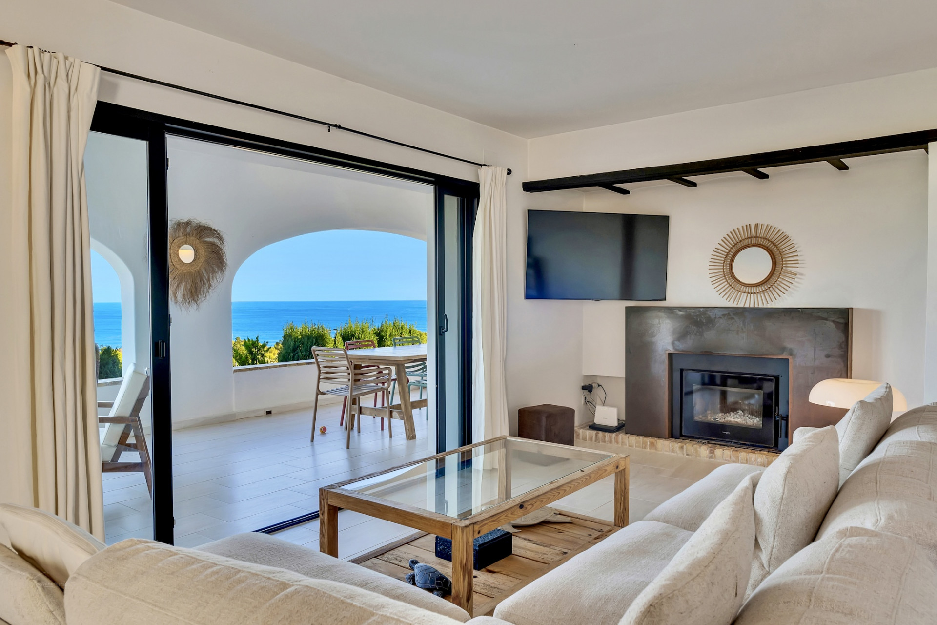 Salón luminoso con sofá claro, mesa de centro de madera y chimenea moderna. Amplios ventanales se abren a una terraza con vistas panorámicas al mar. Salón luminoso con sofá claro, mesa de centro de madera y chimenea moderna. Amplios ventanales se abren a una terraza con vistas panorámicas al mar.